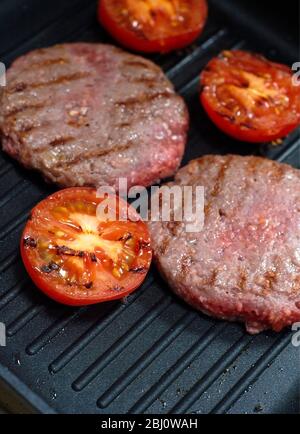 Beefburgers und halbierte Tomaten werden auf Antihaft-Grillpfanne gegrillt - halb gekocht, noch rosa - Stockfoto