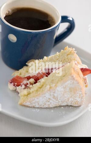 Stück Biskuitkuchen mit Puderzucker bestäubt und mit Erdbeeren und Sahne auf Teller mit kleinen blau-weiß gefleckten Becher schwarzen Kaffees gefüllt - Stockfoto