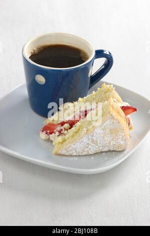 Ein Stück Erdbeerschwamm auf einem weißen Teller mit einem kleinen blau-weiß gefleckten Becher schwarzen Kaffees - Stockfoto