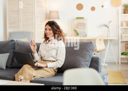 Junge Erwachsene Frau sitzt auf dem Sofa zu Hause Teilnahme an Videokonferenz, grüßt ihre Kollegen Stockfoto