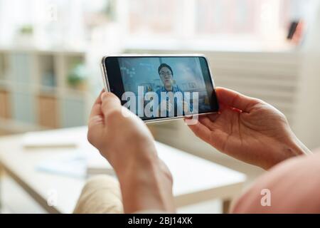 Unkenntlich erkennbare Person, die beim Online-Meeting auf dem Smartphone einen asiatischen Mann beim Sprechen beobachtet, horizontale Nahaufnahme Stockfoto