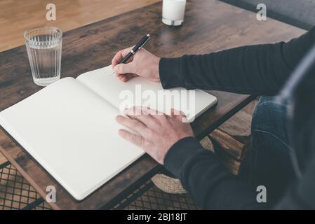 Rückansicht des Mannes, der am Couchtisch sitzt und in Tagebuch oder Notizbuch schreibt Stockfoto