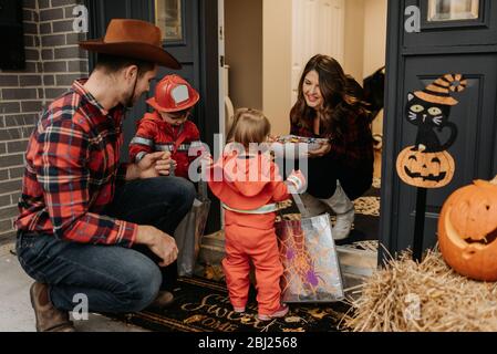 Eine Familie, die sich für Halloween-Trick oder Behandlung verkleidet hat und Süßigkeiten von einer Frau an ihrer Tür sammelt. Stockfoto