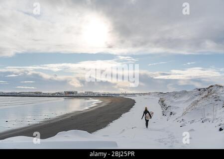 Rückansicht einer Frau, die einen Neoprenanzug trägt und ein Surfbrett hält, das an einem verschneiten Strand entlang läuft. Stockfoto