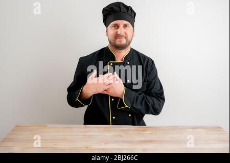 Portrait des jungen männlichen Kochs in schwarzer Uniform hält ihre Hände auf der Brust nahe ihrem Herzen posiert auf einem weißen isolierten Hintergrund Stockfoto