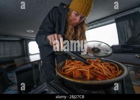 Eine Frau in einem Wohnmobil rührt Essen in einem Topf trägt einen wolligen Hut. Winter Road Trip. Stockfoto