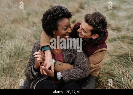 Eine schwarze Frau und ein weißer Mann umarmen sich und lächeln, sitzen in einem Feld von Gras. Stockfoto