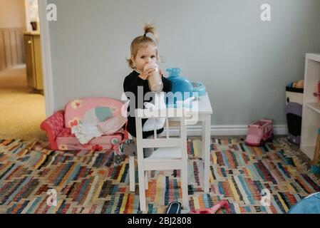 Ein Kind kniet auf einem kleinen weißen Stuhl an einem weißen Tisch und trinkt aus einer Flasche. Stockfoto