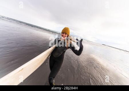 Eine Frau, die einen Neoprenanzug trägt und ein Surfbrett trägt, geht über einen schwarzen Strand mit dem Meer im Hintergrund. Stockfoto