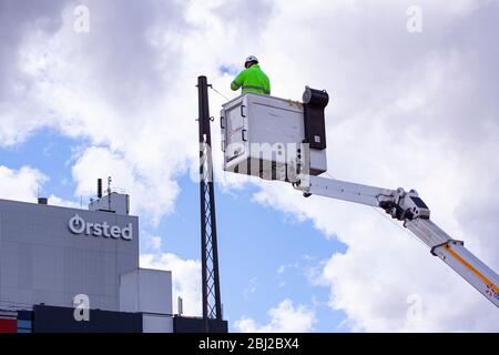 Lineworker arbeiten an Stromleitungen in Kraneimer hoch in der Luft aus der Luft Aufzug. Ein Ørsted Kraftwerk im Hintergrund. Sonniger Tag mit blauem Himmel. C Stockfoto