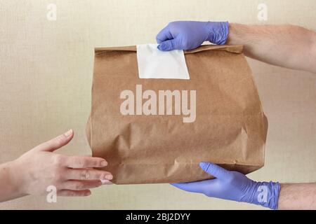 Männliche Hände in blauem Latex Handschuh gibt Papiertüte an weibliche Hände. Nahaufnahme der männlichen Hände in Handschuhen halten braunen Bastelpapierbeutel mit leeren weißen Aufkleber. D Stockfoto