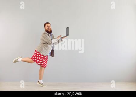 Lustige dicke Mann in einer Jacke arbeitet mit einem Laptop beim Tanzen auf einem grauen Hintergrund. Stockfoto