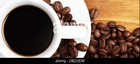 Eine weiße Tasse Kaffee auf einer Untertasse, verstreute Körner der gleichen braunen Farbe auf einem Tisch aus strukturierten Holzbrettern. In der Ecke des Bildes befindet sich das Stockfoto