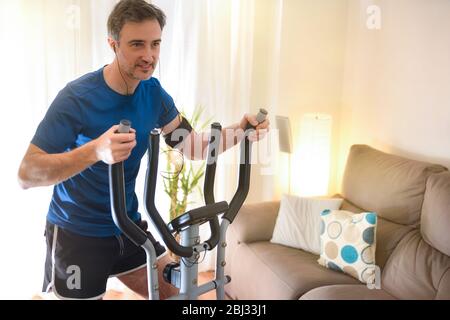 Lächelnder Mann, der im Wohnzimmer elliptischen Fahrradsport macht und Musik mit Kopfhörern und Telefon hört Stockfoto