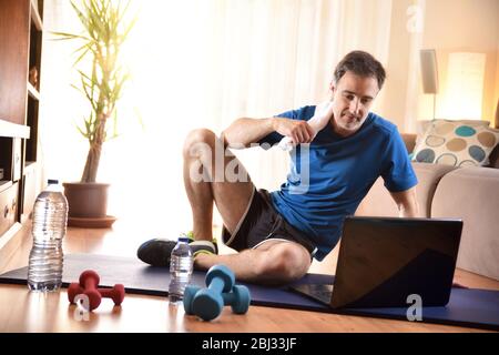 Mann, der Sportbekleidung auf einer Matte in seinem Wohnzimmer trägt und Videos auf einem Laptop anschaut. Stockfoto