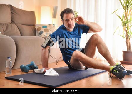 Mann, der sich nach dem Sport ausgeruht hat und Musik mit Kopfhörern auf dem Telefon auf einer Matte in seinem Wohnzimmer hört Stockfoto