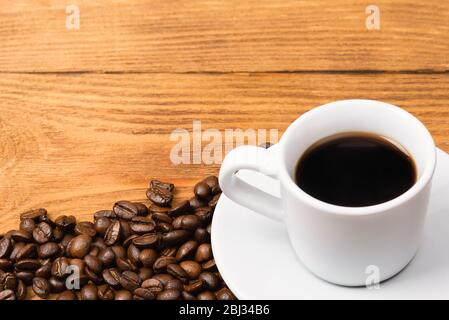 Frisch gemachter Kaffee in einer weißen Tasse auf einer weißen Untertasse, umgeben von verstreuten Kaffeebohnen auf einem Holztisch. Frisch gerösteter Kaffee. Platz für einen Inscri Stockfoto