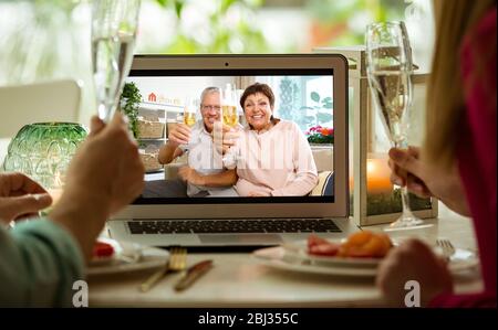 Mann und Frau sitzen am Esstisch, essen, trinken Champagner und haben Videoanruf mit älteren Eltern auf dem Laptop. Zu Hause bleiben Stockfoto