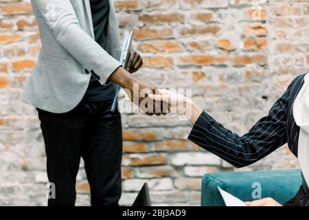 Geschäftskonzept des Handschüttelns. Zugeschnittenes Bild von Händeschütteln nach Vereinbarung zwischen Geschäftsfrau in schwarzer Jacke und dunkelhäutigen Geschäftsmann in Stockfoto
