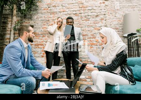 Multiethnische Geschäftsleute auf dem Meeting. Muslimische Frau und kaukasischen Mann Lesung Projekt berichtet sitzen auf den Sofas, während blonde Frau und afrikaner Stockfoto