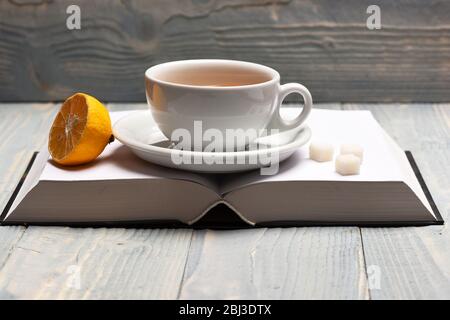 Teetasse auf leeren Buchseiten, die gemütliche Komposition machen. Tasse grüner Tee mit Zitronenscheibe. Warmes Getränk und Home Relax Konzept. Heißes Getränk mit Zuckerwürfeln auf offenem Buch auf Holzhintergrund gelegt Stockfoto