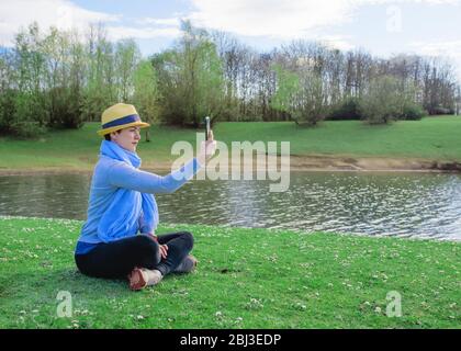 Frau in einem grauen Top, Strohhut, schwarze Hose, blauer Schal, die ein Foto am Telefon und lächelnd, sitzt auf einer Wiese in der Nähe des Wassers an sonnigen Frühling Stockfoto