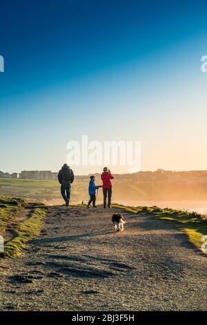 Eine Familie und ihr Hund spazieren auf dem Küstenweg, während die Sonne über die Fistral Bay in Newquay in Cornwall untergeht. Stockfoto