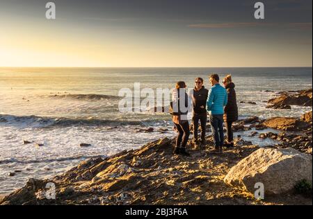 Eine Gruppe von Freunden, die sich unterhalten und auf Felsen mit Blick auf Little Fistral in Newquay in Cornwall stehen. Stockfoto