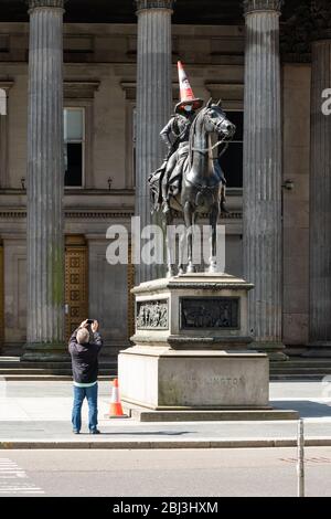 Glasgow, Schottland, Großbritannien. April 2020. Glasgow während der Sperrung des Coronavirus. Ein Mann nimmt ein Foto der legendären Statue des Duke of Wellington mit einer Gesichtsmaske auf, um zu Mittag zu essen. Quelle: Kay Roxby/Alamy Live News Stockfoto