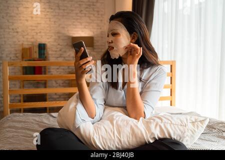 Porträt von jungen asiatischen Mädchen Anwendung Gesichtsmaske auf ihr Gesicht, während mit Computer-Laptop im Schlafzimmer. Frauen Beauty und Gesichtspflege Konzept. Stockfoto