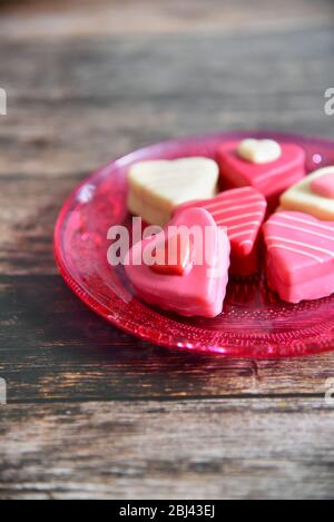 Herzförmige, farbenfrohe Pralinen auf einem rosa Glasteller mit Ornamentmuster. Dunkler Holztisch Hintergrund. Liebe und Grußkarte Konzept. Stockfoto