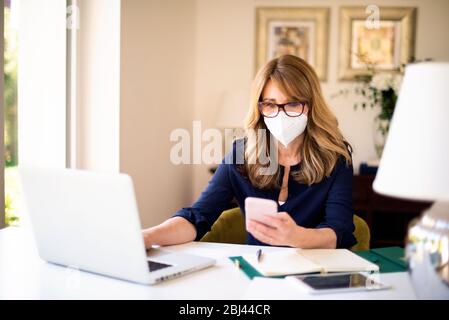Porträtaufnahme einer Frau mittleren Alters, die während der Arbeit an ihrem Notebook zu Hause eine Gesichtsmaske trägt und während der Coronavirus-Pandemie Textnachrichten übergibt. Stockfoto