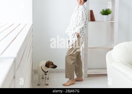 Ruhe und Gemütlichkeit. Schöne junge Frau mit Hund stand neben dem Fenster zu Hause, Nahaufnahme. Stockfoto