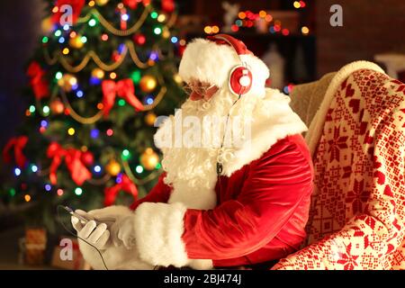 Weihnachtsmann sitzt mit Kopfhörern in bequemen Schaukelstuhl zu Hause sitzen Stockfoto