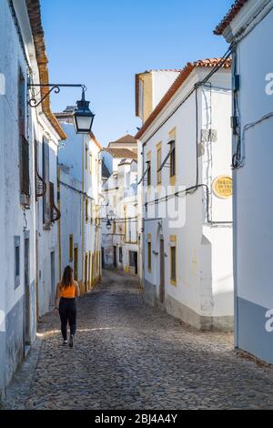 Junge Frau in lässiger Kleidung, die in einer Kopfsteinpflasterstraße durch typische weiße und gelbe Häuser in Evora, Portugal, geht Stockfoto