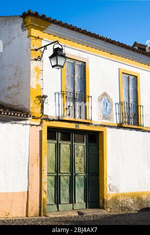 Typisches weiß-gelbes Haus mit Balkon, Laterne, alter Eingangstür und azulejo blauen Fliesen religiöser Kunst in Evora, Portugal Stockfoto