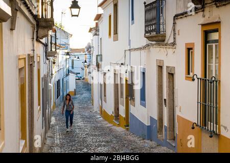 Junge Frau in lässiger Kleidung, die in einer Kopfsteinpflasterstraße durch typische weiße und gelbe Häuser in Evora, Portugal, geht Stockfoto