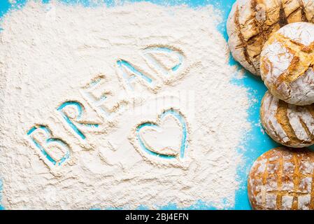 Das Wort Brot schrieb in der Mehlplatte auf einem blauen Tisch. Haufen von runden weißen Broten. Hausgemachtes Sauerteig Brot Stockfoto