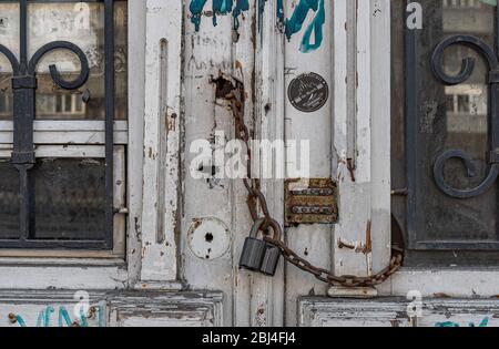 Architektonische Details der grobkörnigen weißen Holztür, die von einem alten rostigen Vorhängeschloss verkettet ist. Kunstvolle geschwungene Gitter in den Türfenstern eines verlassenen Retro-Gebäudes. Stockfoto