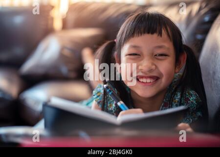 Glücklich kleines Kind Mädchen auf dem Sofa mit dem Lernen von Laptop und Schreiben eines Buches zu Hause, soziale Distanz während der Quarantäne, Online-Bildung Konzept Stockfoto