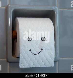 Toilettenpapier-Rolle mit fröhlichen Gesicht in einem Toilettenpapierhalter, grau-blaue Fliesen Stockfoto