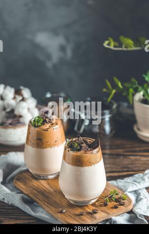 Zwei Gläser Iced Dalgona Coffee, ein trendiger, flauschiger, cremiger Schlagkaffee, dekoriert mit Thymian und dunkler Schokolade. Dunkler rustikaler Stil. Stockfoto