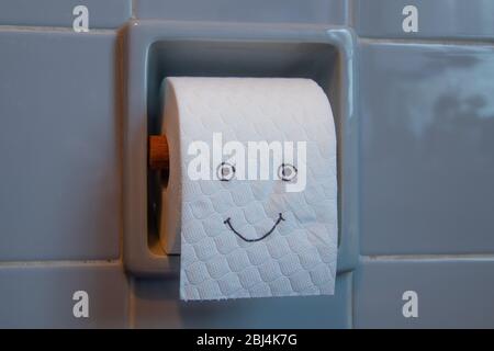 Toilettenpapier-Rolle mit fröhlichen Gesicht in einem Toilettenpapierhalter, grau-blaue Fliesen Stockfoto