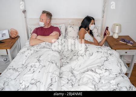 Paar im Bett trägt Gesichtsmasken nach Streit während der Selbstisolierung während in Lockdown, Frau, die ihr Telefon überprüft Stockfoto