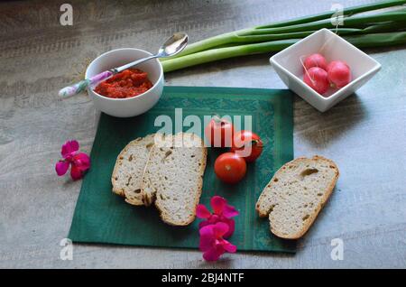 Gesunde Frühstücksindicenthaltsstoffe auf einem Holztisch: Handwerkliches Brot, Kirschtomaten, Frühlingszwiebeln, ajvar in einer weißen Schüssel, Rettich, Rucola Stockfoto