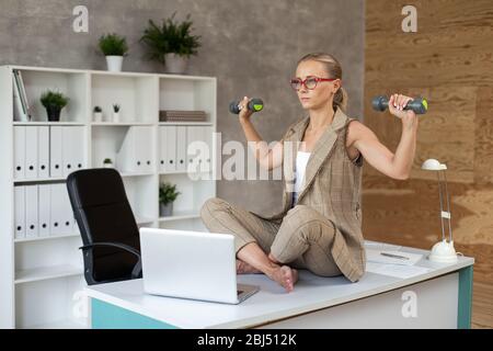 Gut aussehende Geschäftsfrau sitzt in Lotushaltung mit zwei Hanteln in den Händen. Stockfoto