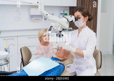 Ein Zahnarzt untersucht die Mundhöhle einer Frau mit Hilfe eines speziellen medizinischen Geräts, während ihre kleine Tochter sie beobachtet. Stockfoto