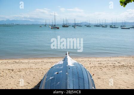 Auf dem Kopf stehendes Holzboot im Schatten eines Baumes am Strand mit mehreren Segelbooten und Booten, die im Hintergrund festgemacht sind. Bucht mit ruhigem Meer und keine Wellen n Stockfoto