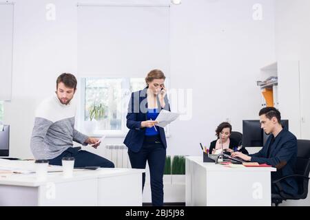 Büromitarbeiter in einem offenen Raum, verschiedene Dinge tun, Kollegen chatten, Projekte diskutieren, an PCs arbeiten, etwas Papierkram erledigen. Coworking-Kontra Stockfoto