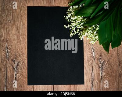 Blumenstrauß Maiglöckchen und leeres schwarzes Blatt auf rustikalem Holztisch von oben, schöne Vintage-Karte, Draufsicht, Platz für Text Stockfoto